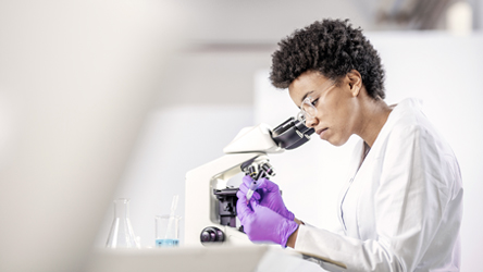 Mulher em laboratório, devidamente equipada, usa microscópio para desenvolvimento de análises cientificas.