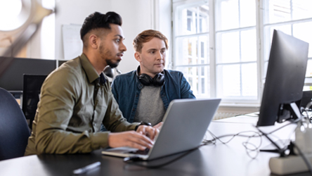Dois homens jovens, analisam informação através dos seus computadores em ambiente de escritório