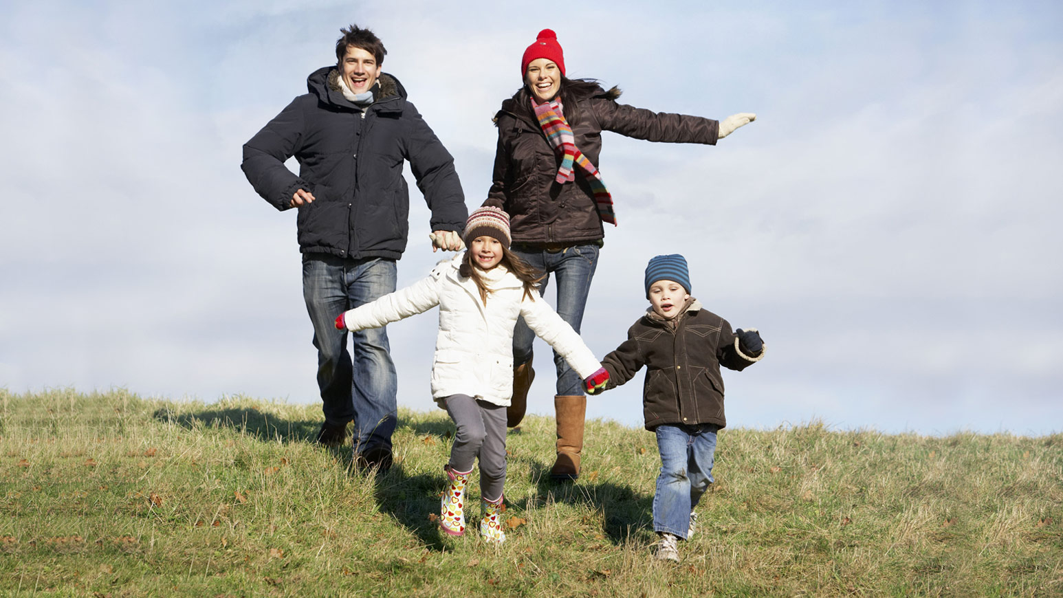 Quatro elementos de uma possível família correndo num campo relvado num dia frio. Os pais de mão dada atrás e os filhos à frente.