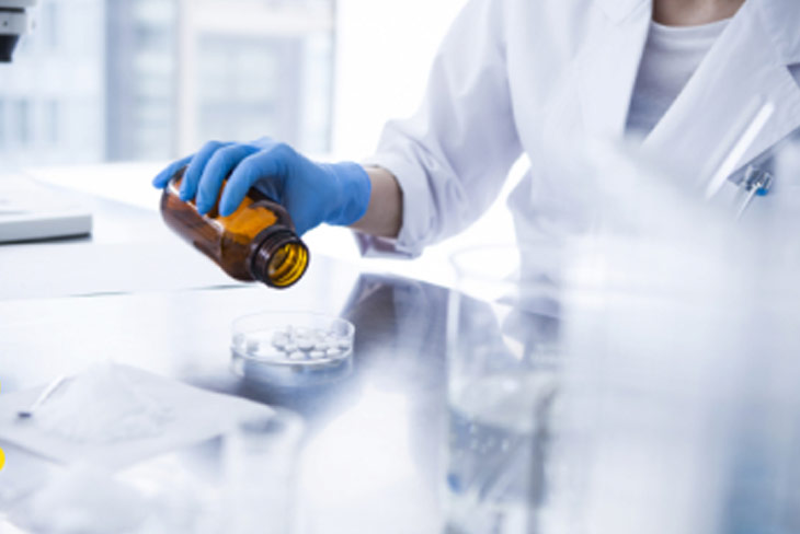 Fotografia de um laboratório, com um profissional de bata branca e luvas de latex a manusear um frasco de um medicamento, depositando os comprimidos num recipiente de laboratório.