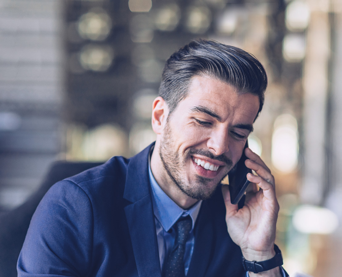 Um homem sorridente, de fato, participa numa chamada telefónica usando um telemóvel.